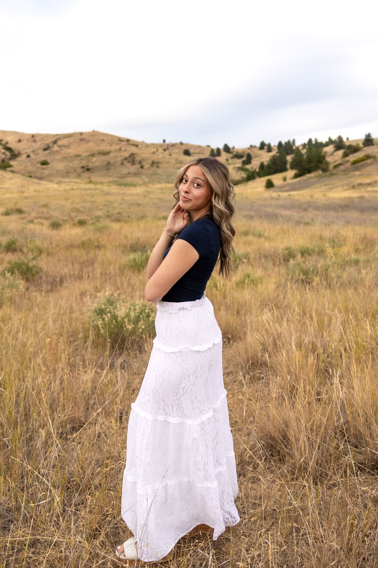 A woman in a white and navy dress poses gracefully in a golden autumn field against rolling hills.