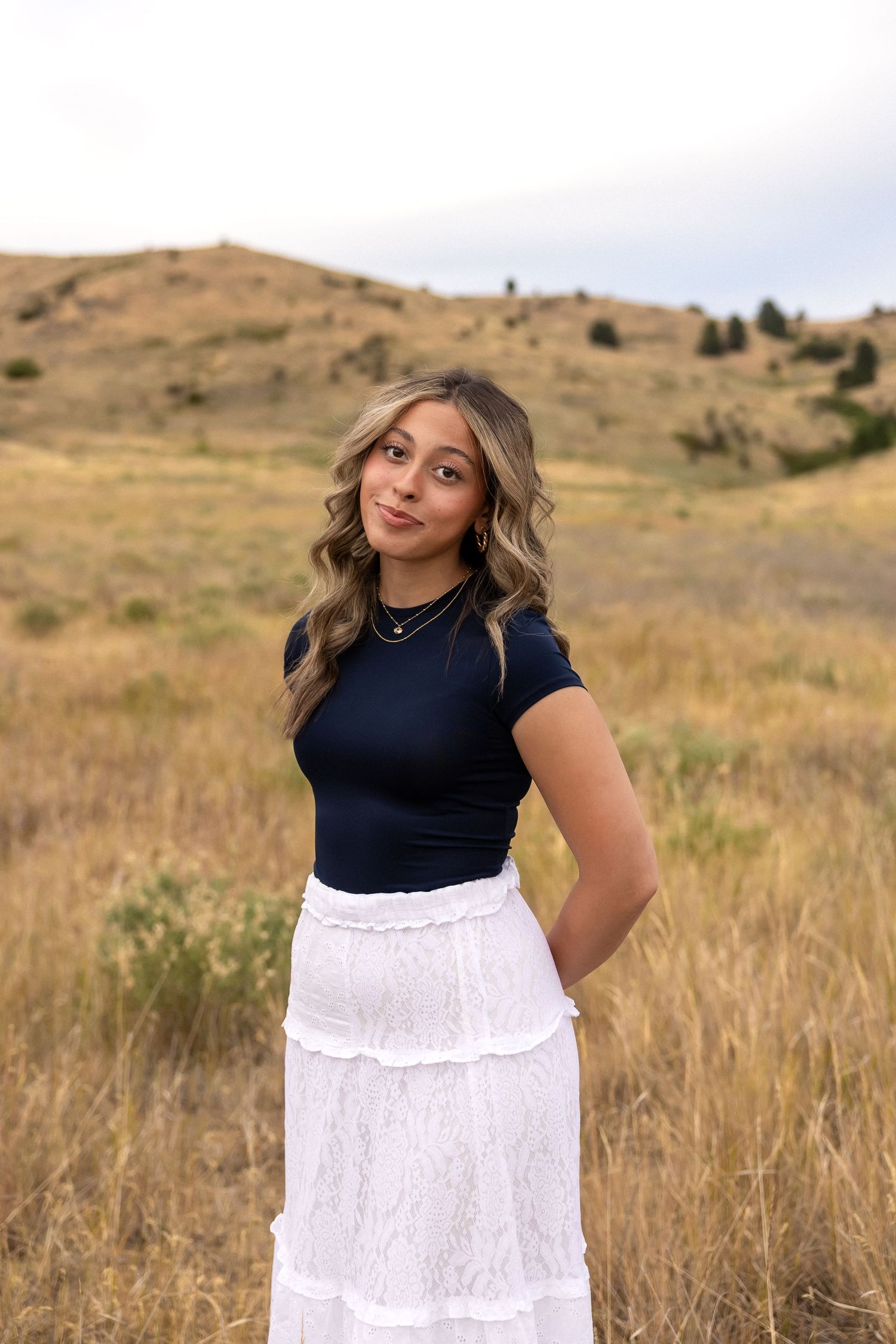 Person in a navy top and white tiered maxi skirt poses in front of rolling hills during golden hour.