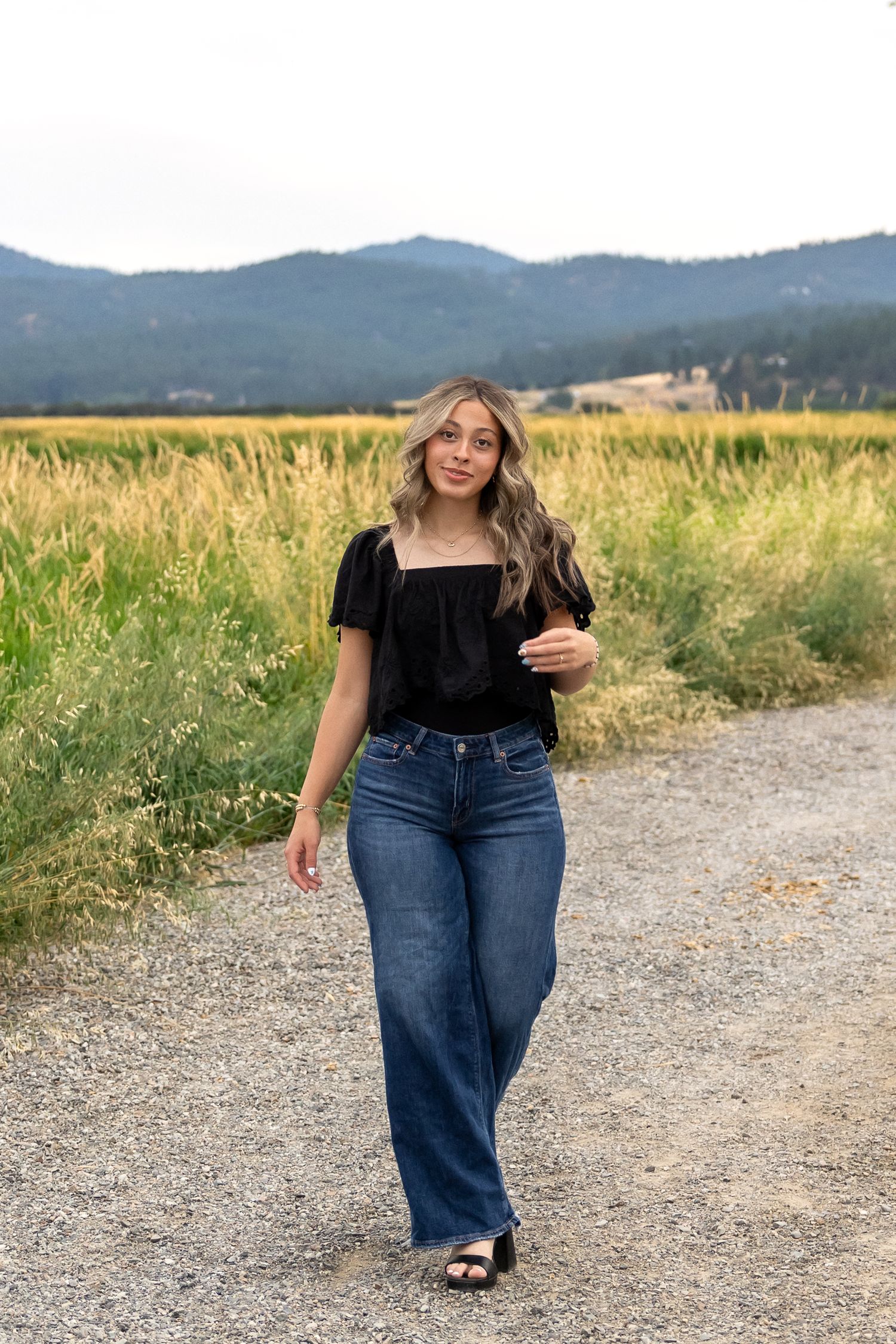 A casual summer outfit featuring blue jeans and a black top against a mountain landscape backdrop.