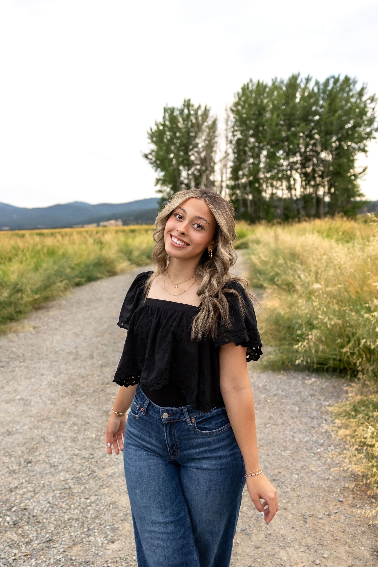 A person in a black top and jeans poses joyfully on a dirt path with mountains and trees in the background.
