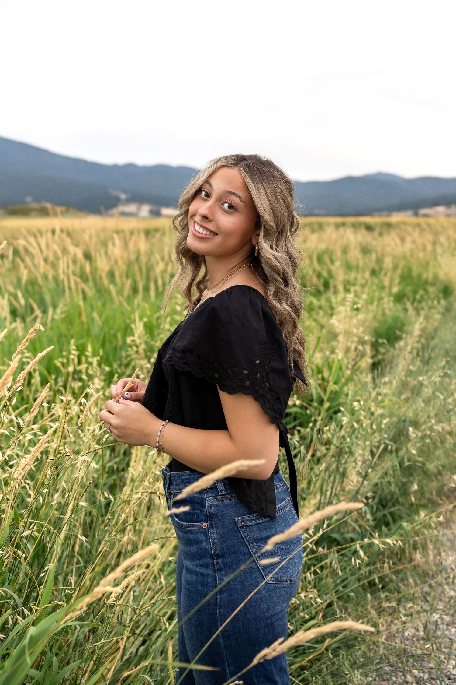 Person in black top and denim overalls poses playfully in a field of tall grass at sunset with mountains in background.