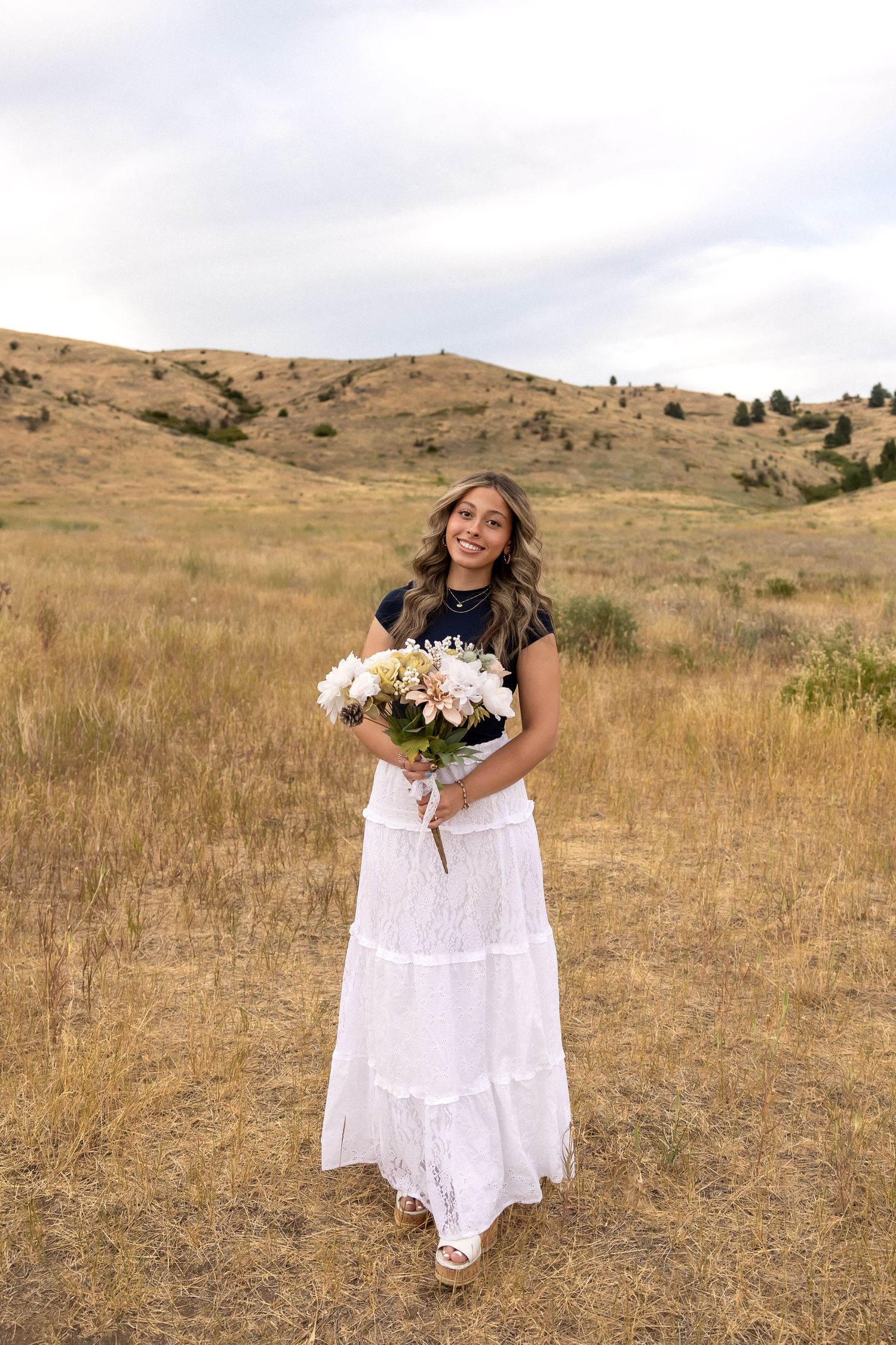 A woman in a white lace dress holds a bouquet while standing in a golden field with rolling hills in the background.