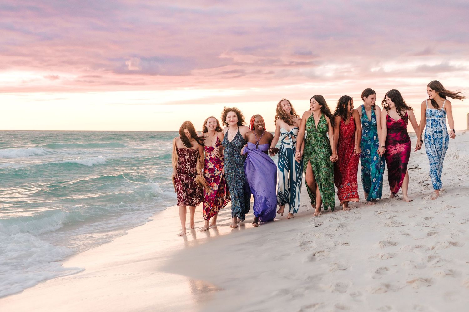 A group of friends in colorful dresses walk together along a beach at sunset with waves crashing beside them.