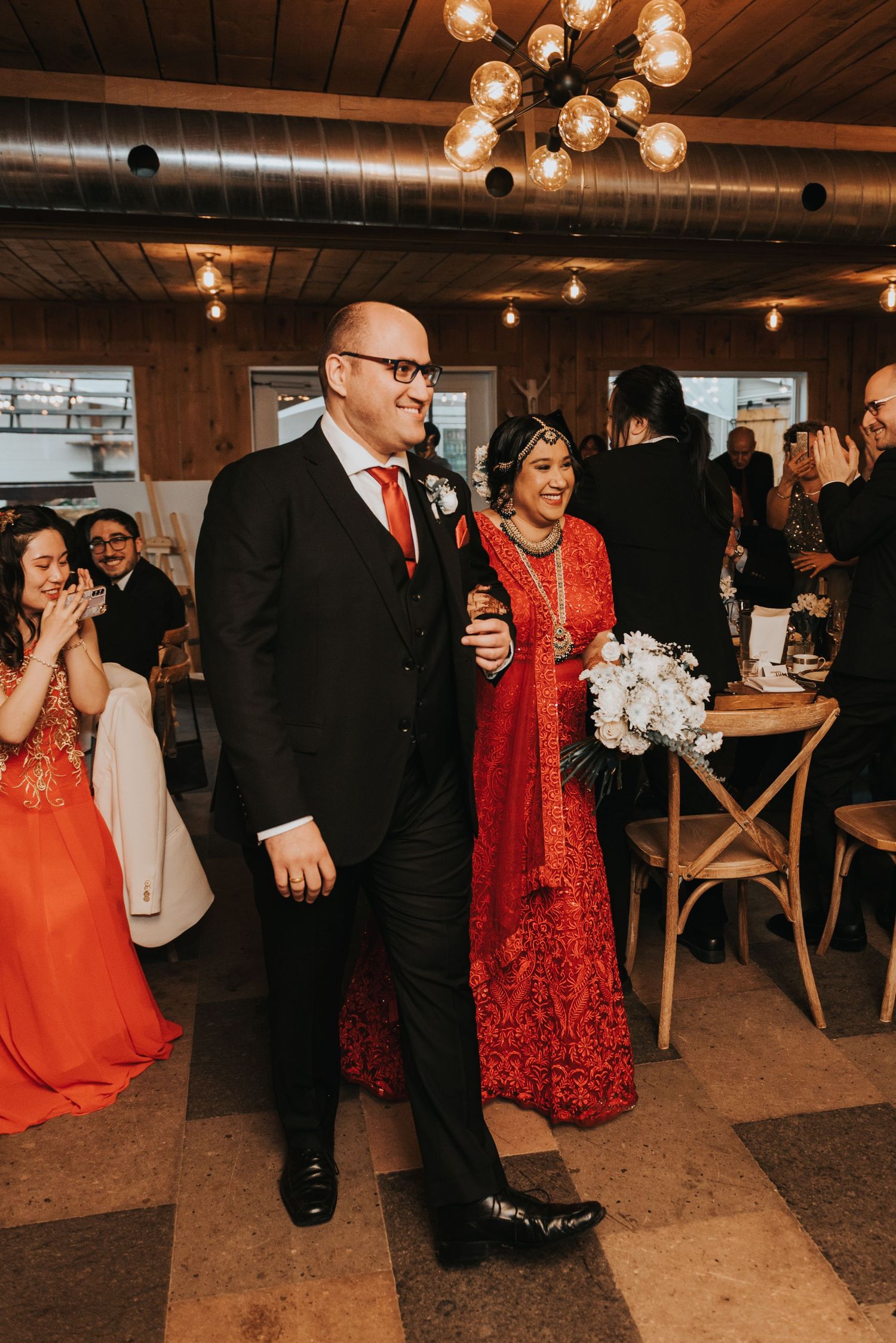Wedding guests dance and celebrate on a wooden floor under warm lighting at an indoor reception venue.