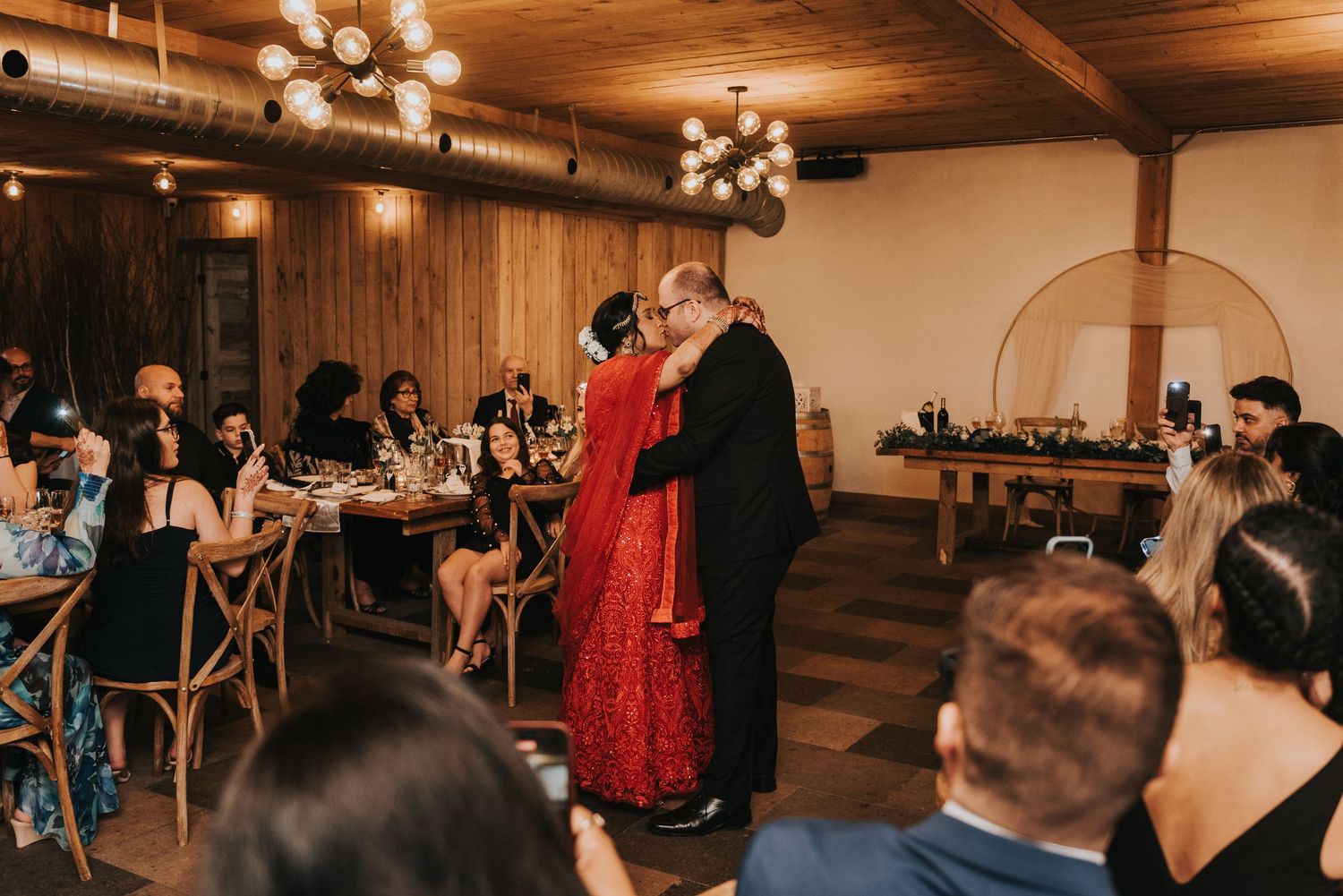Wedding guests dance and socialize in a rustic wooden venue with warm lighting and elegant decor.