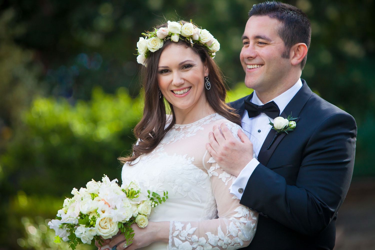 A happy couple on their wedding day share a joyful moment outdoors with white floral bouquet and flower crown.