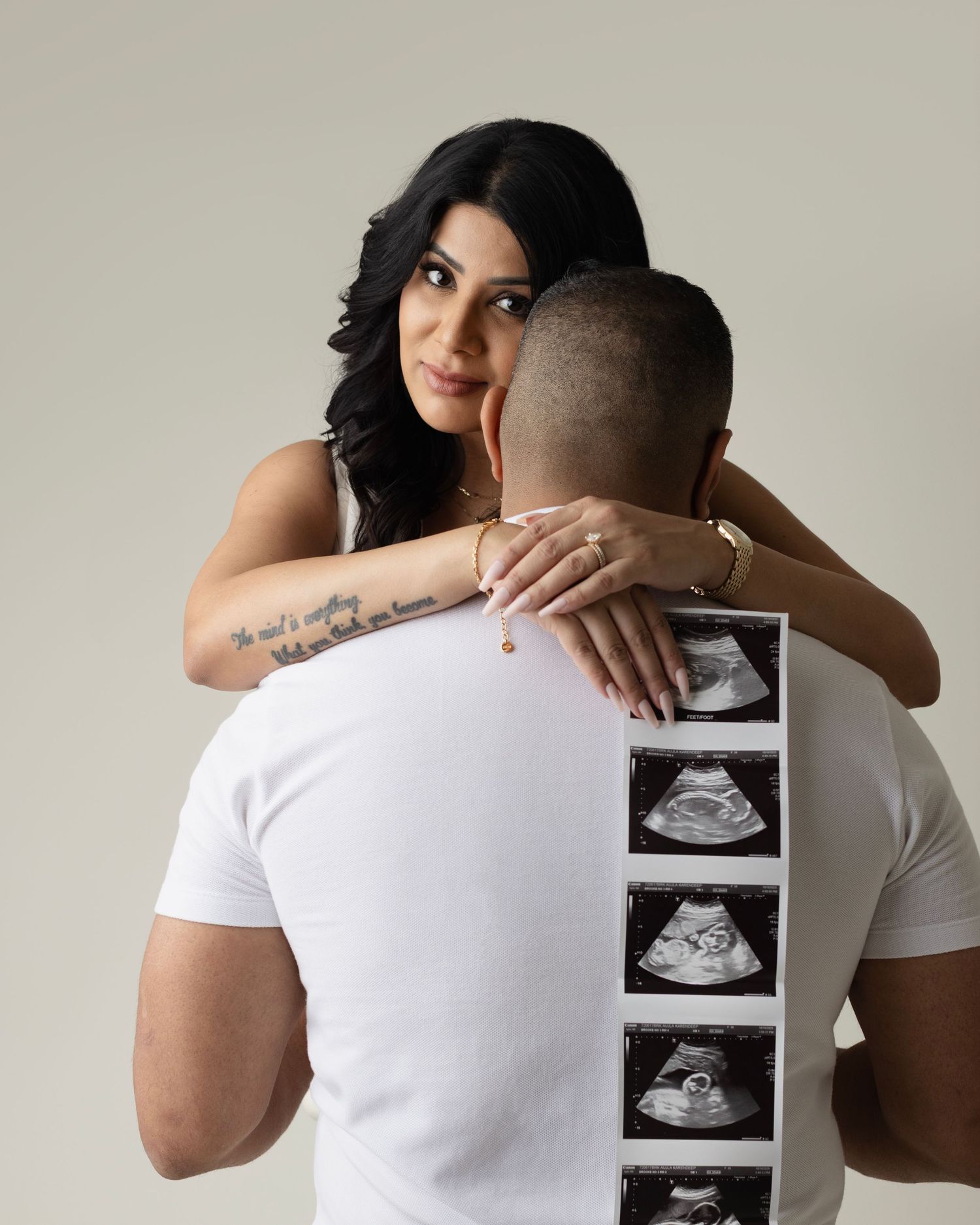 A couple embraces while holding up an ultrasound photo against a light gray background in a pregnancy announcement series.