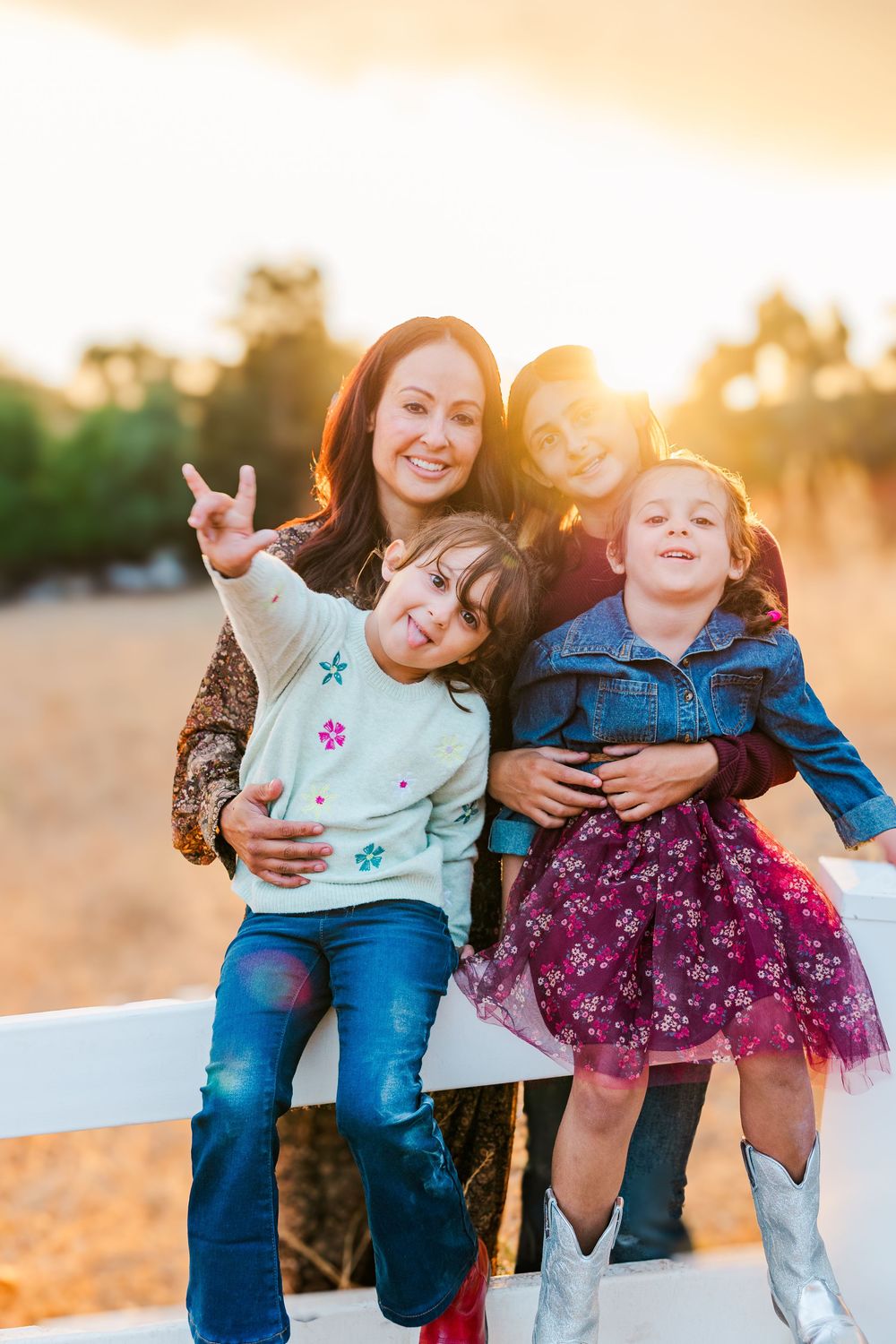 Family members sitting on a white fence during golden hour sunset with children in denim and purple outfits.