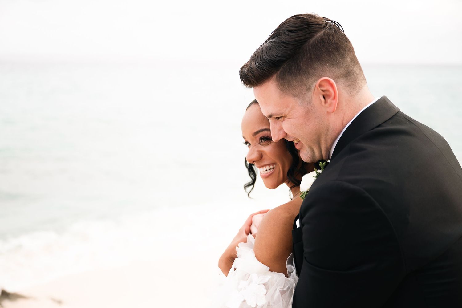 A joyful couple in formal wedding attire celebrates their special day with lively poses against an oceanfront backdrop.