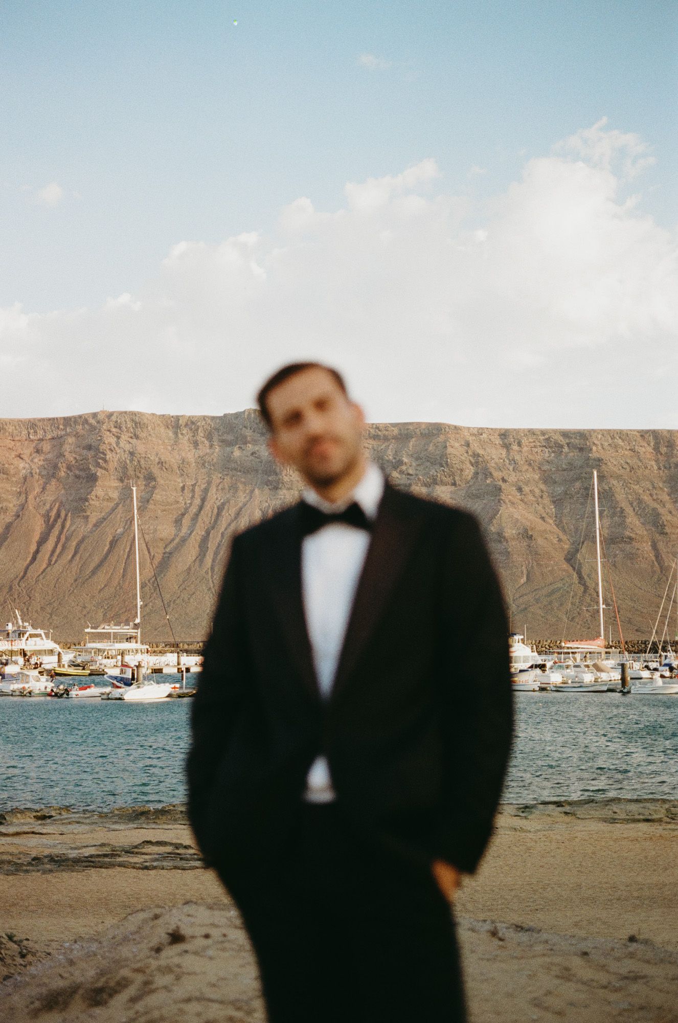 Scenic beach landscape with boats anchored near cliffs and a person in formal attire standing on sandy shore.