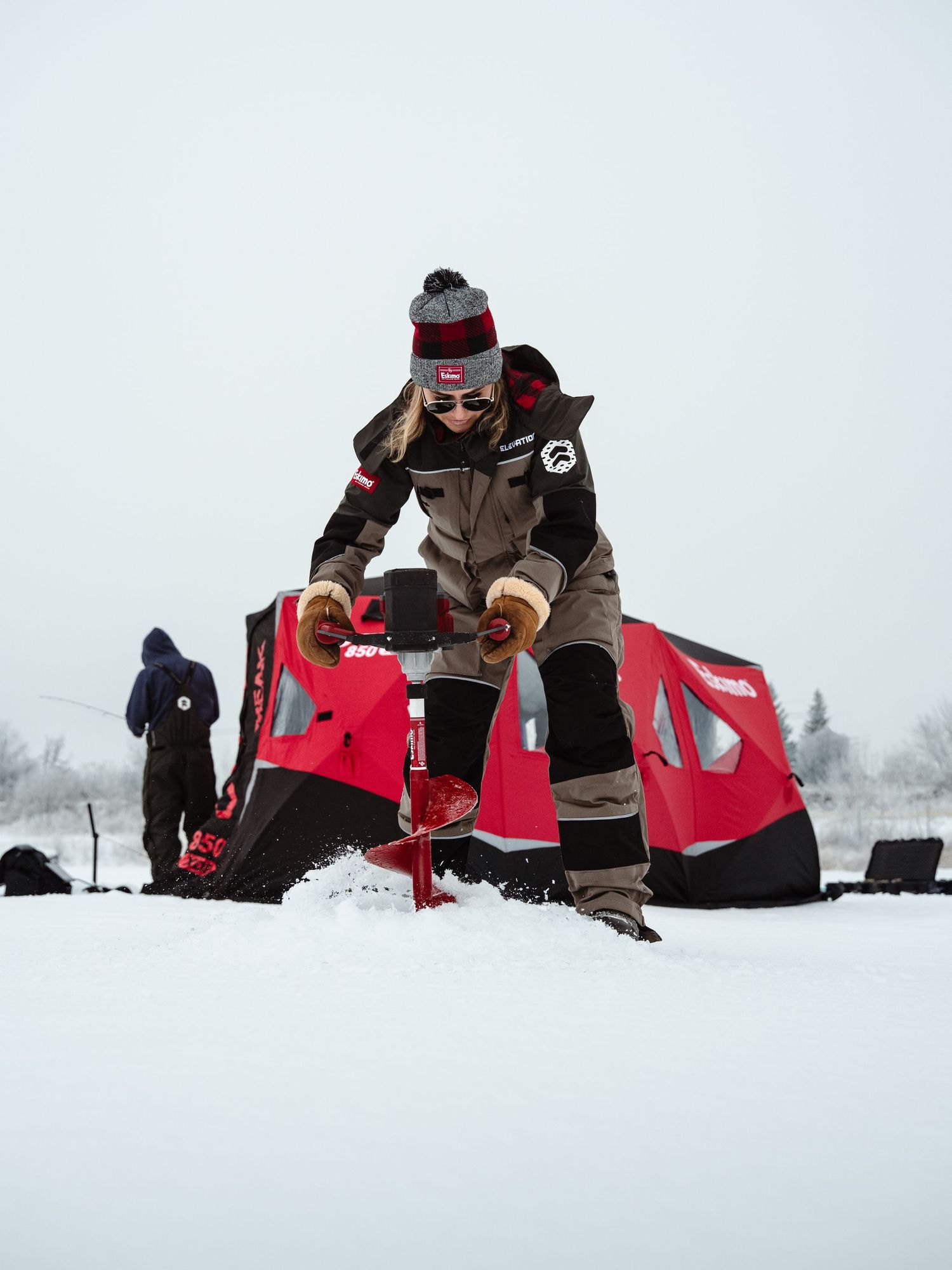 Ice fishers in red and brown winter gear setting up equipment on frozen lake.