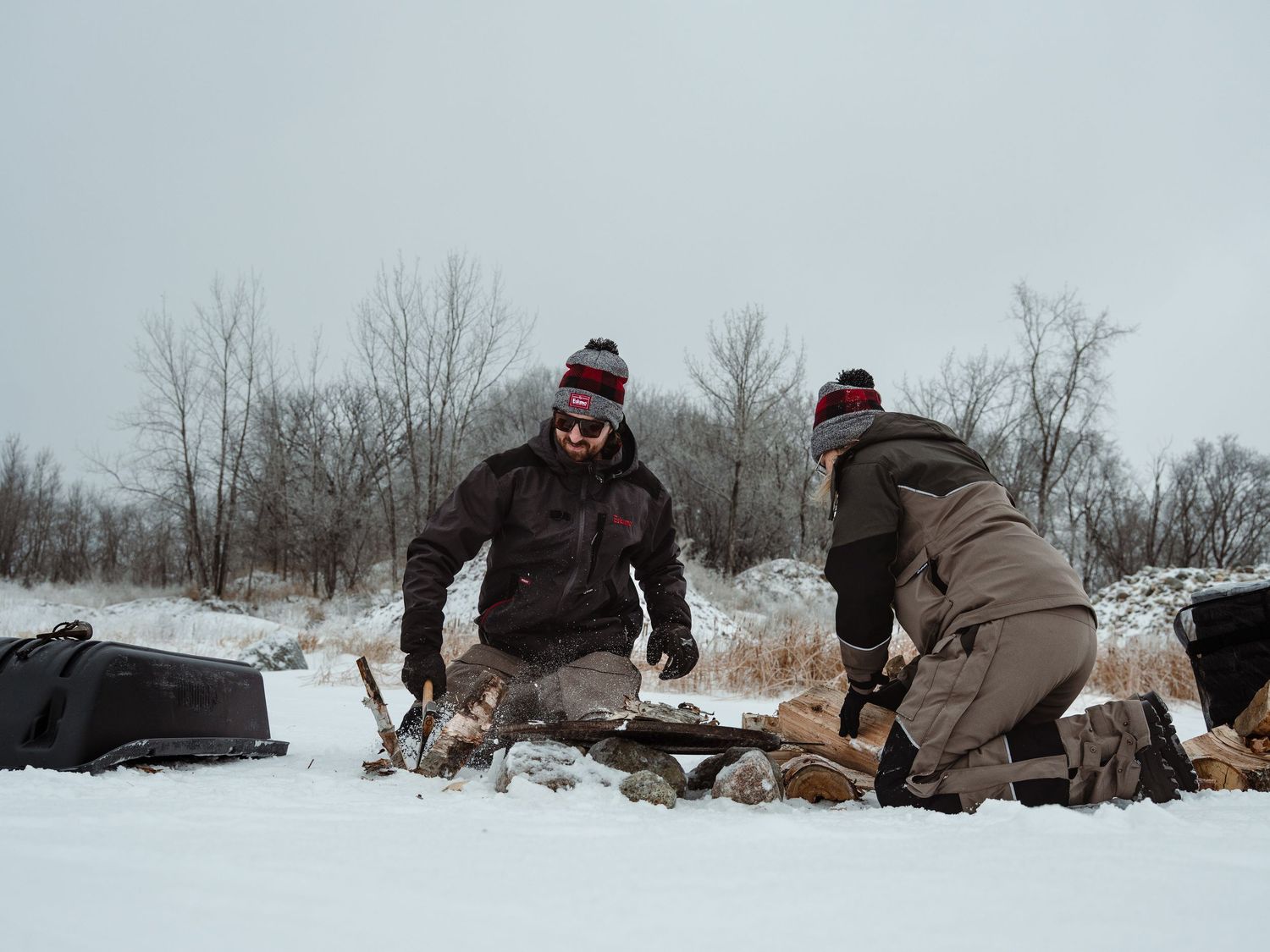 Ice fishers preparing equipment on snowy ground with bare trees in background.