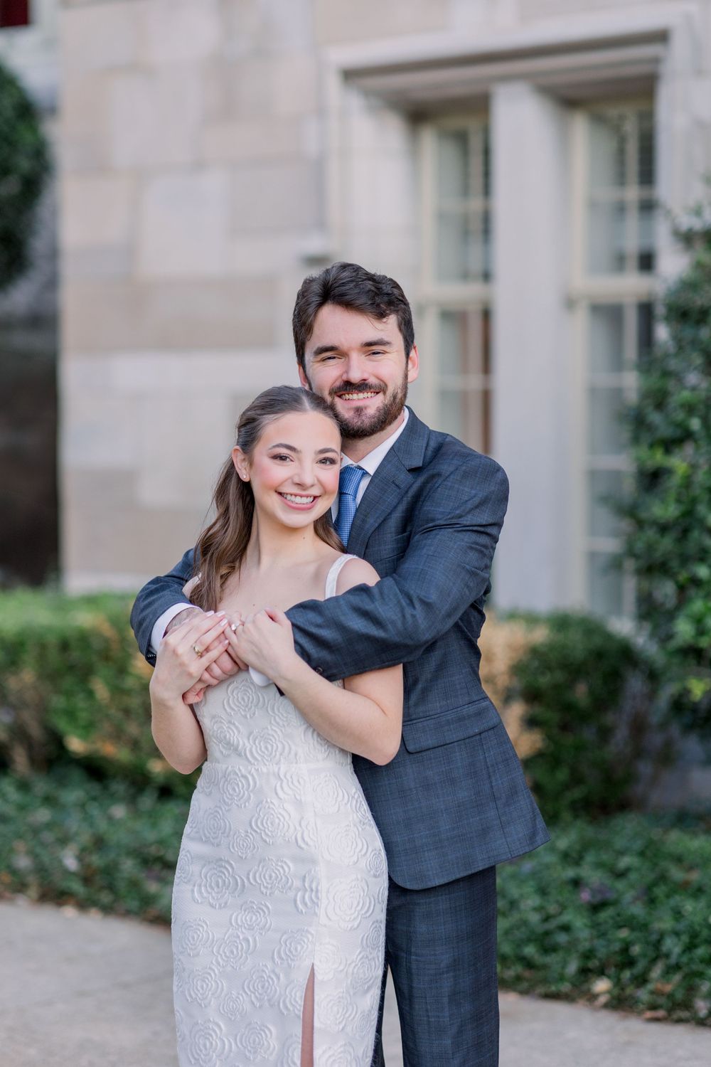 Couple shares tender moment in front of elegant stone building with white trim windows.