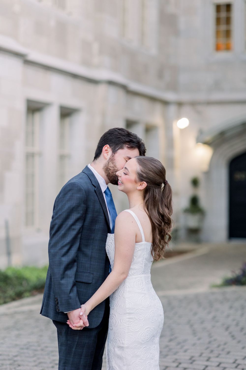Couple embraces and kisses on cobblestone path by stone architecture.