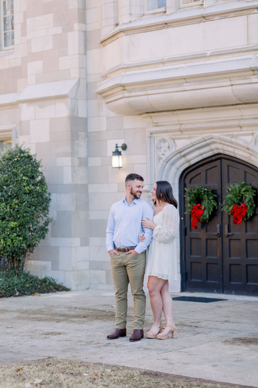 Couple poses together outside stone building decorated with holiday wreaths.