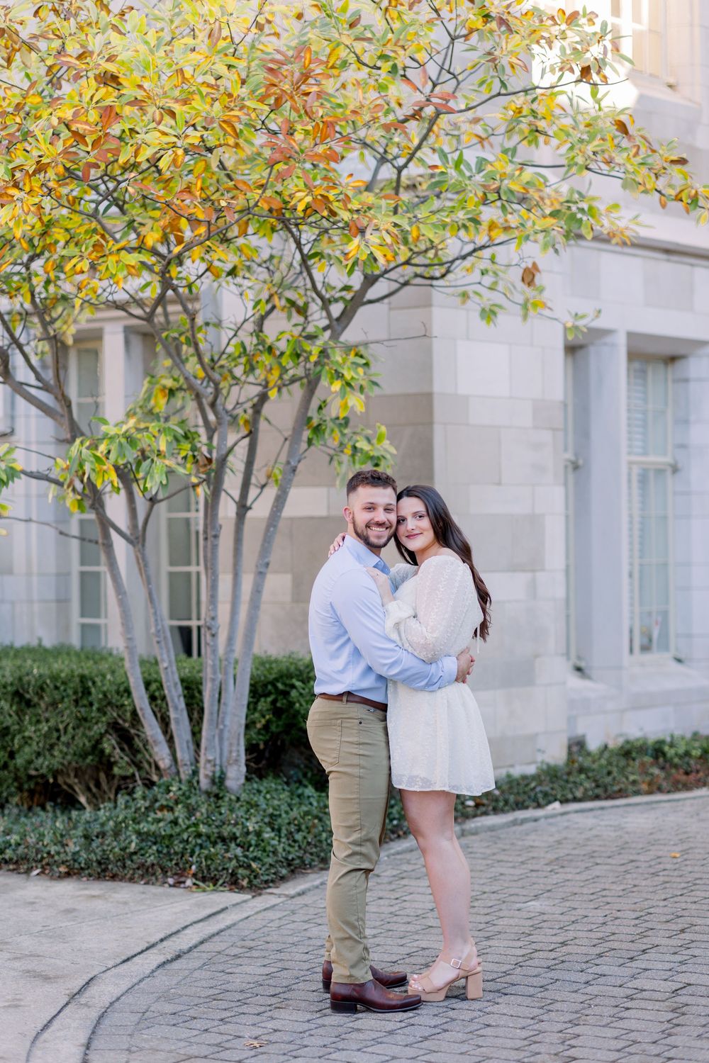 Couple embraces under fall foliage by stone building with cobblestone path.