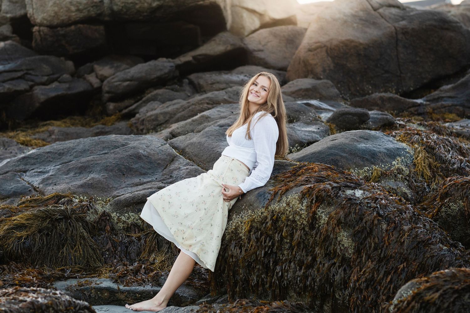 Gloucester, North Shore senior photographer. Long hair blonde girl wearing yellow skirt leans on dark coastal rocks for senior photos.