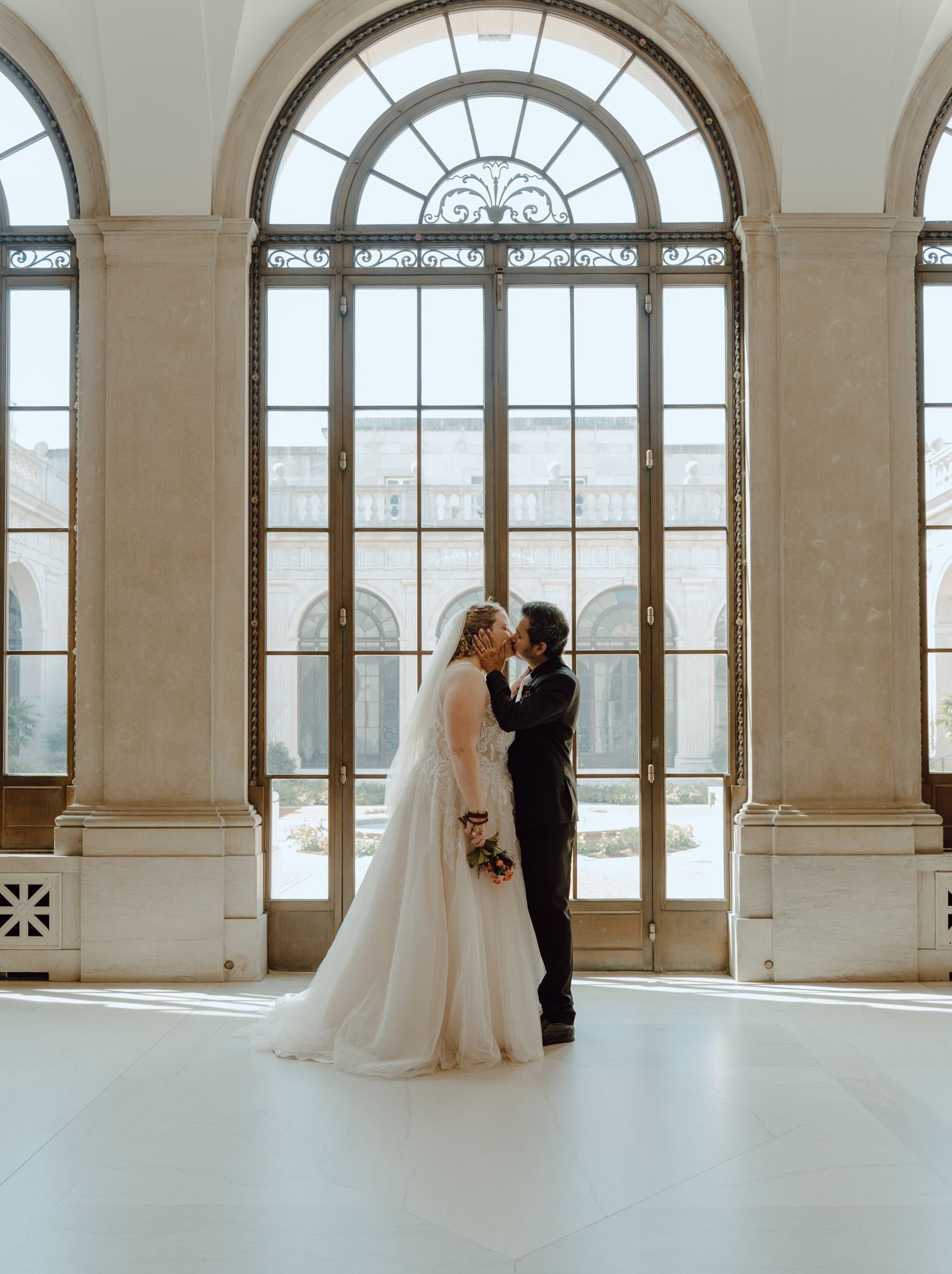 A couple shares an intimate moment in front of grand arched windows in an elegant architectural setting.