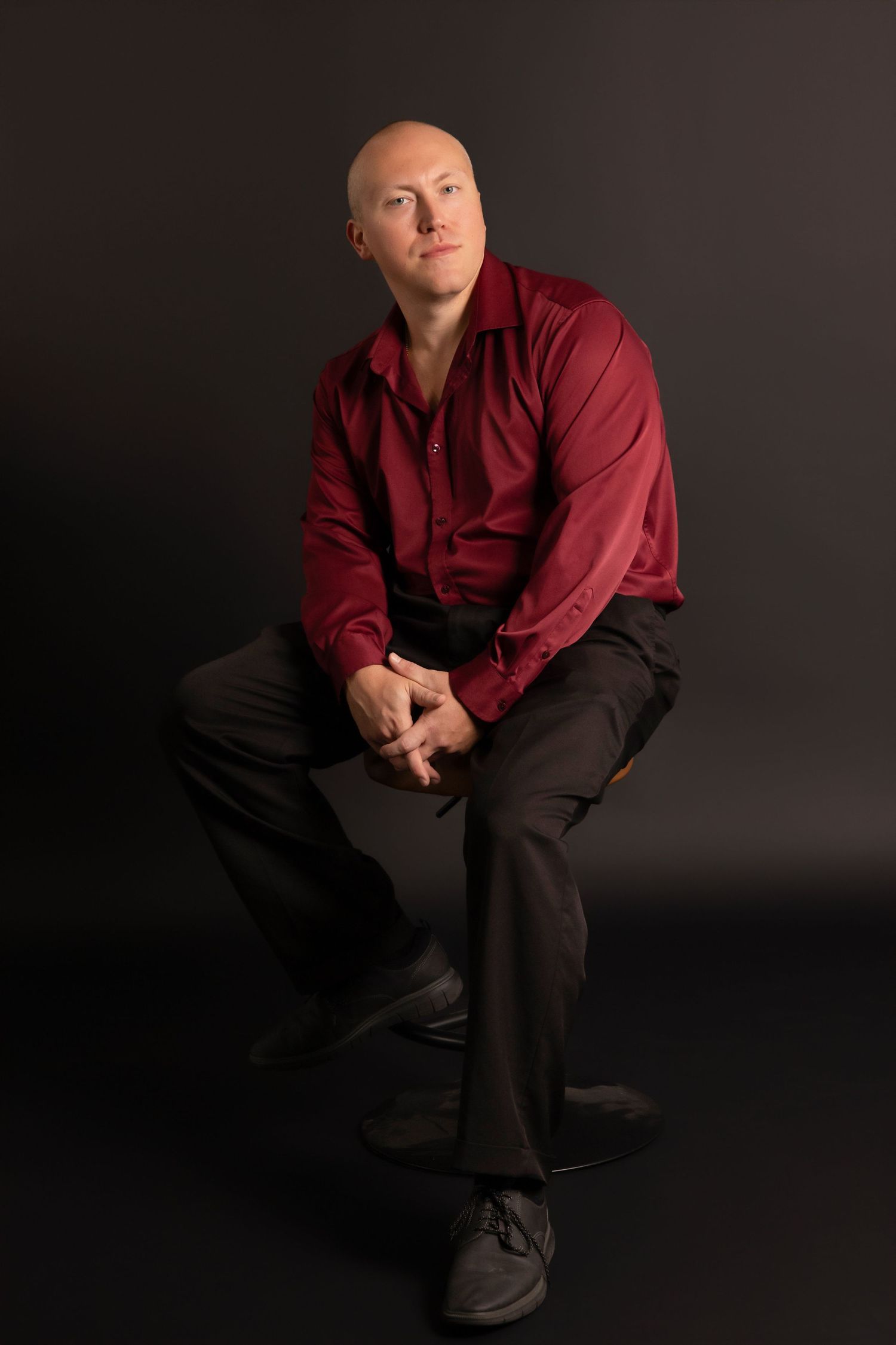 A person in a burgundy shirt and dark pants poses thoughtfully against a black studio backdrop.