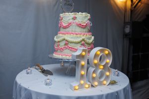 A white wedding cake decorated with pink flowers and gold number 18 lights sits elegantly on a table.