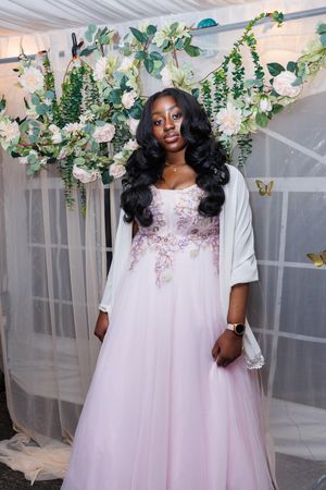 A person wearing a flowing pink tulle gown poses in front of a floral arch with white roses and greenery.