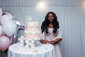 A celebratory scene with a pink and white tiered cake, silver balloons, and white decorative backdrop for a special occasion.