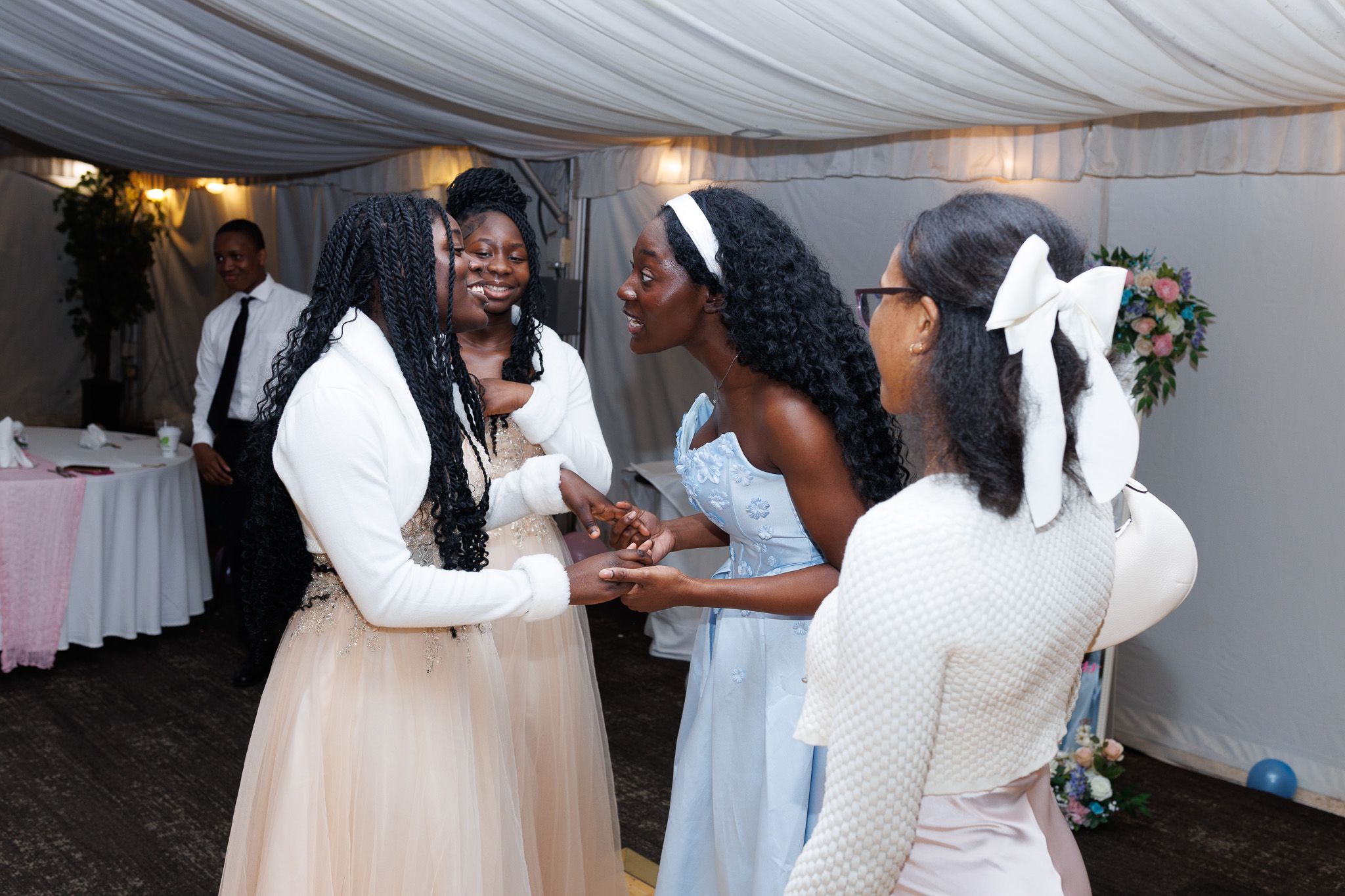 People in white and pastel attire gather and interact at an indoor celebration under draped fabric decor.