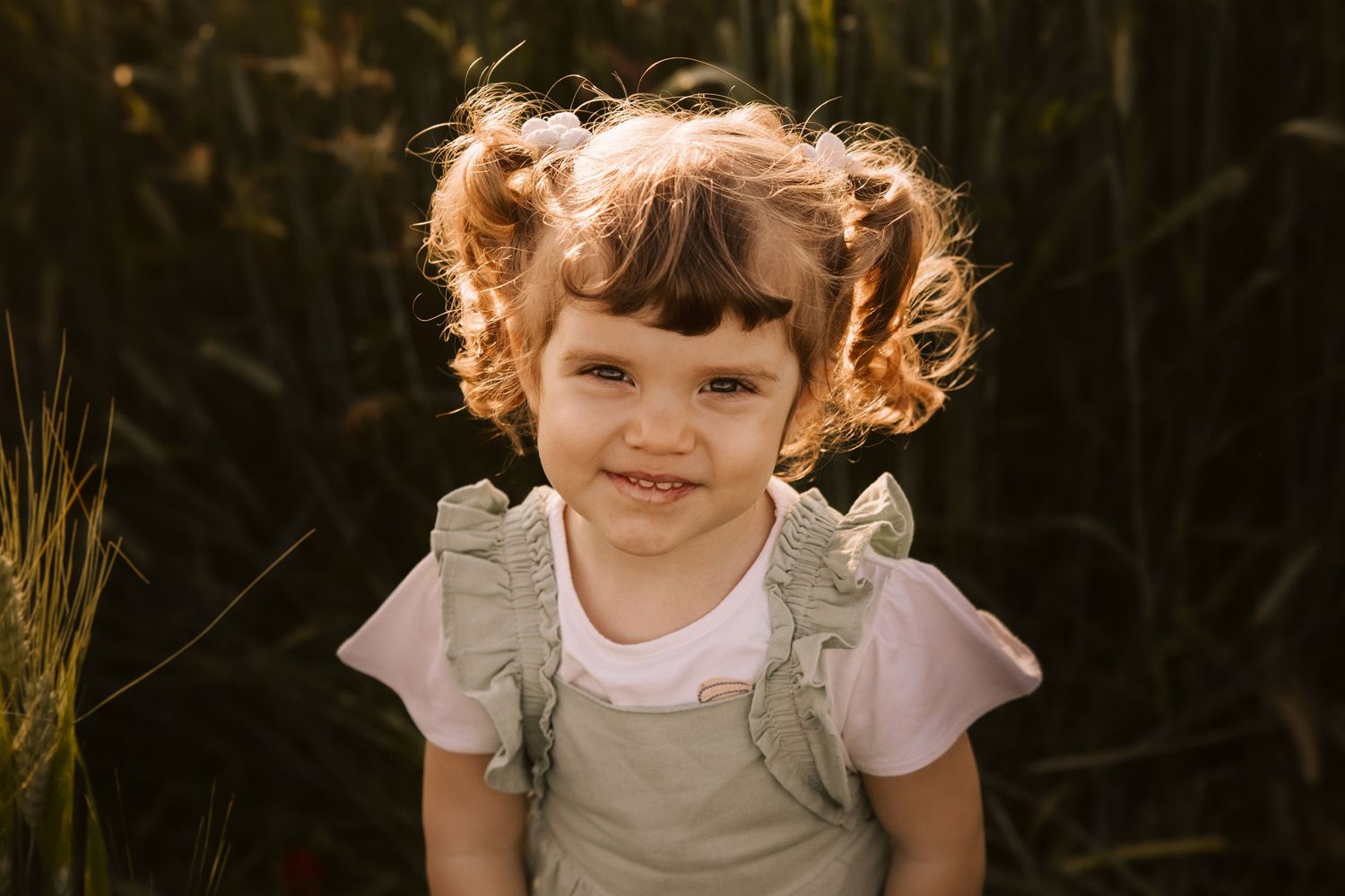 A child with curly pigtails wearing overalls and a white t-shirt smiles while standing in front of tall grass at sunset.