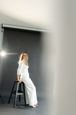 Fashion portrait of woman in white outfit posing on stool in studio with dramatic lighting.