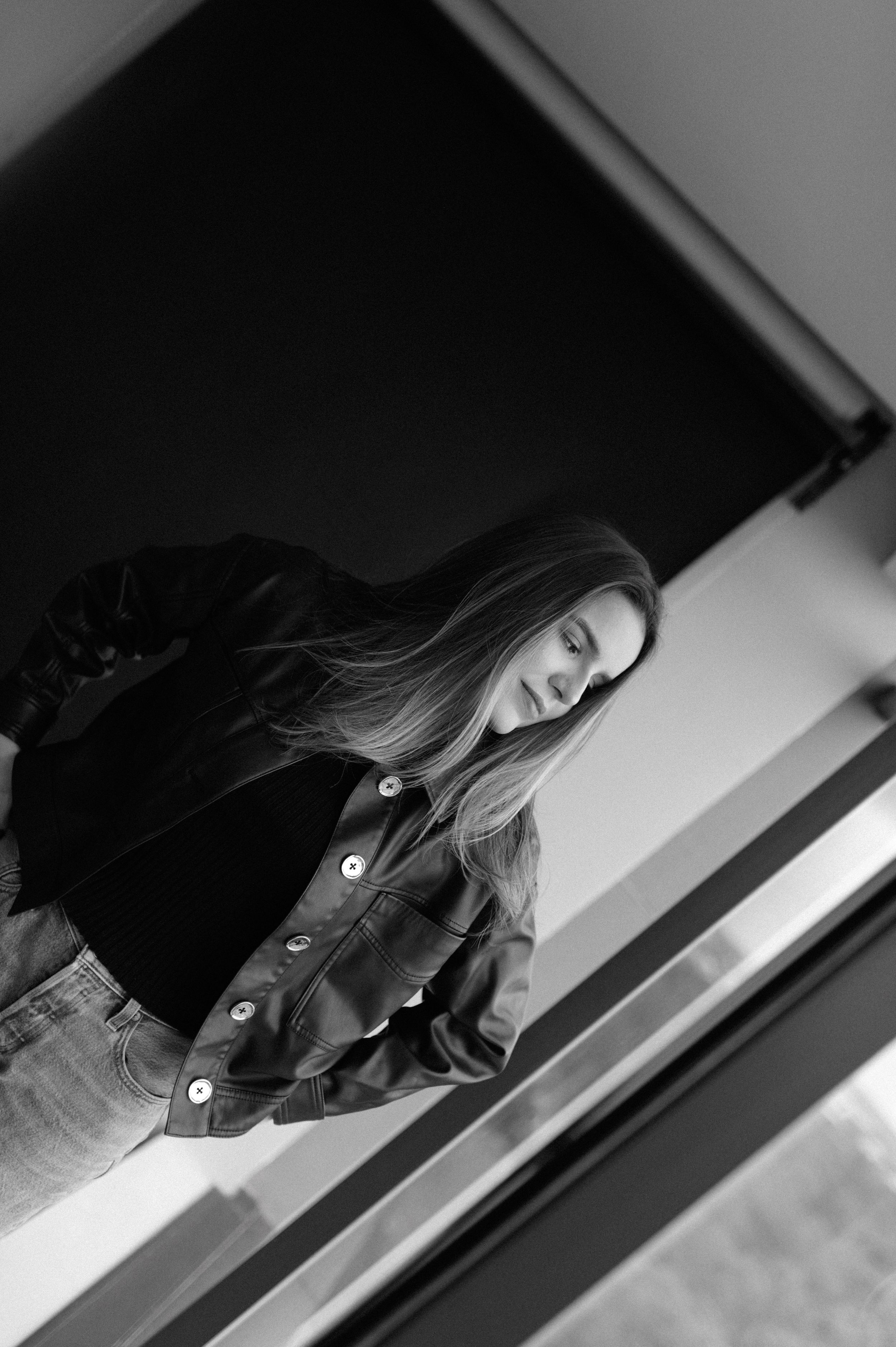 Casual portrait by window showing person in denim and dark button-up shirt with long hair.