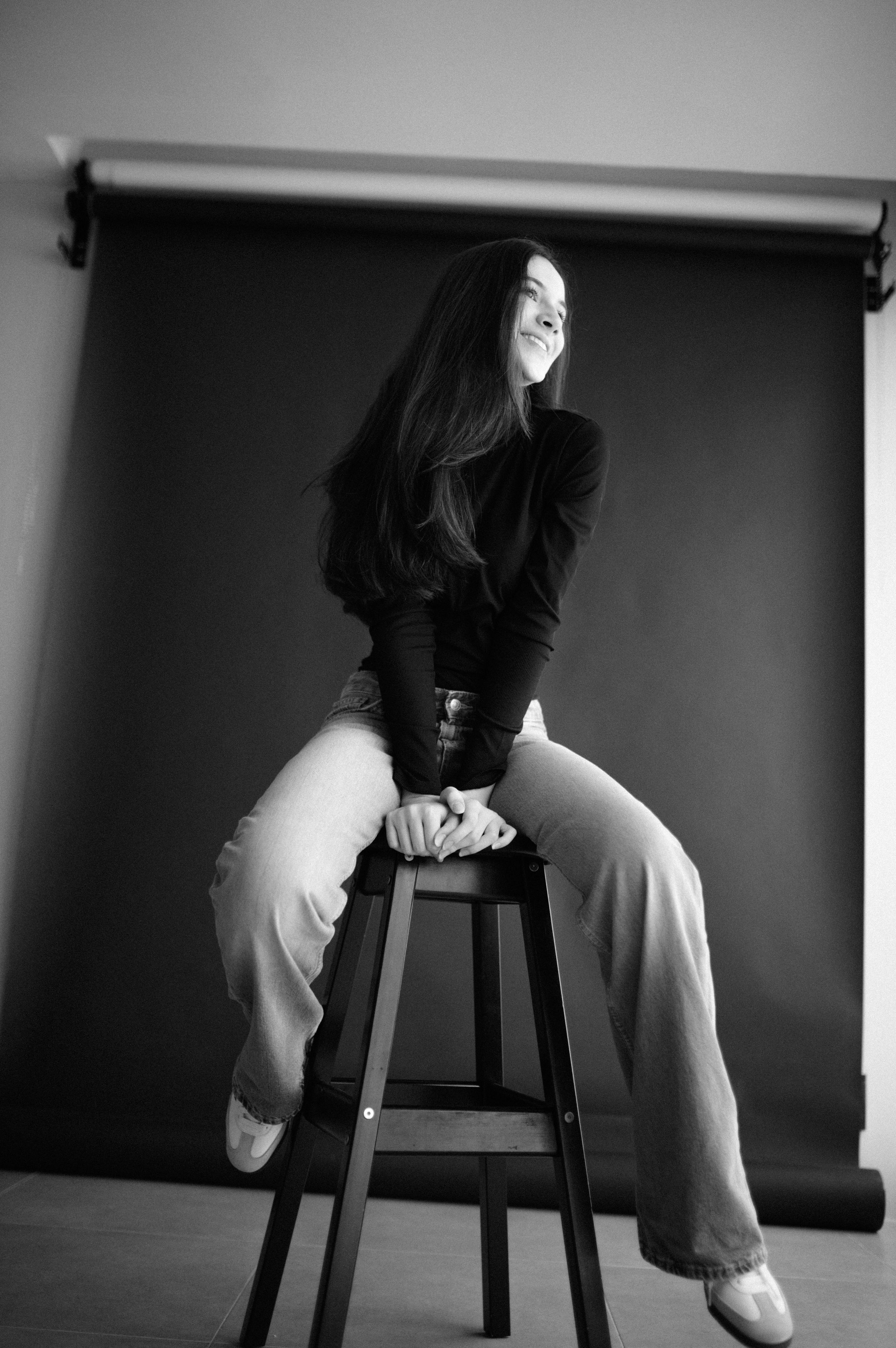 Artistic black and white portrait on stool against studio backdrop showing casual pose.