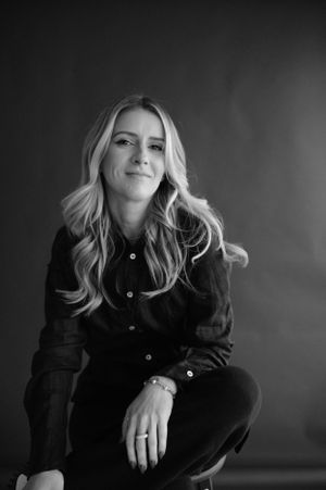 Black and white portrait of a person in dark clothing with wavy blonde hair sitting casually against dark background.