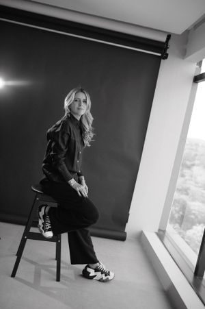 Candid portrait of someone in dark clothing sitting on stool by window in modern setting.