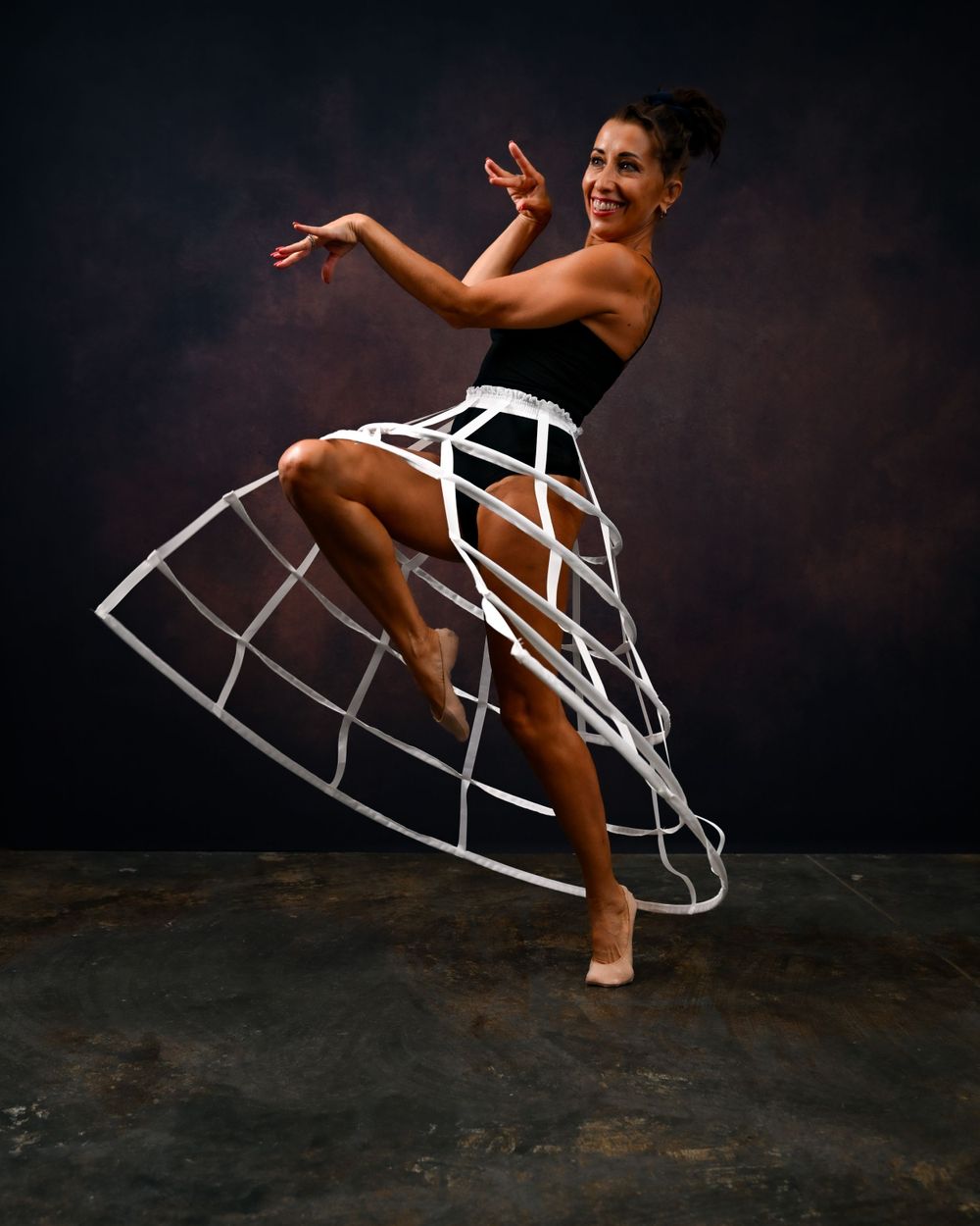 Dancer in structured white cage skirt poses expressively against dark backdrop.
