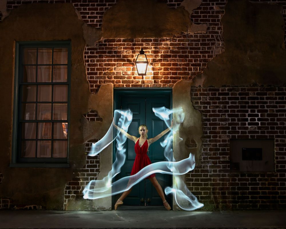 Dancer in red dress creates light trails through movement against brick wall at night.