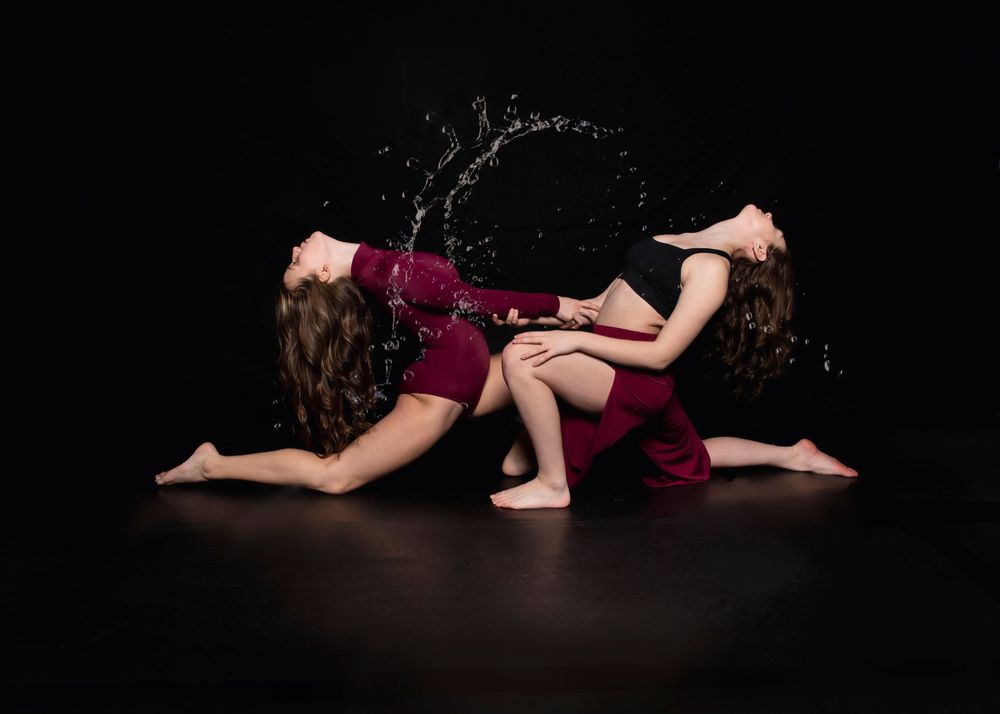 Two dancers in burgundy dresses create a dynamic pose splash water while performing on dark stage floor.