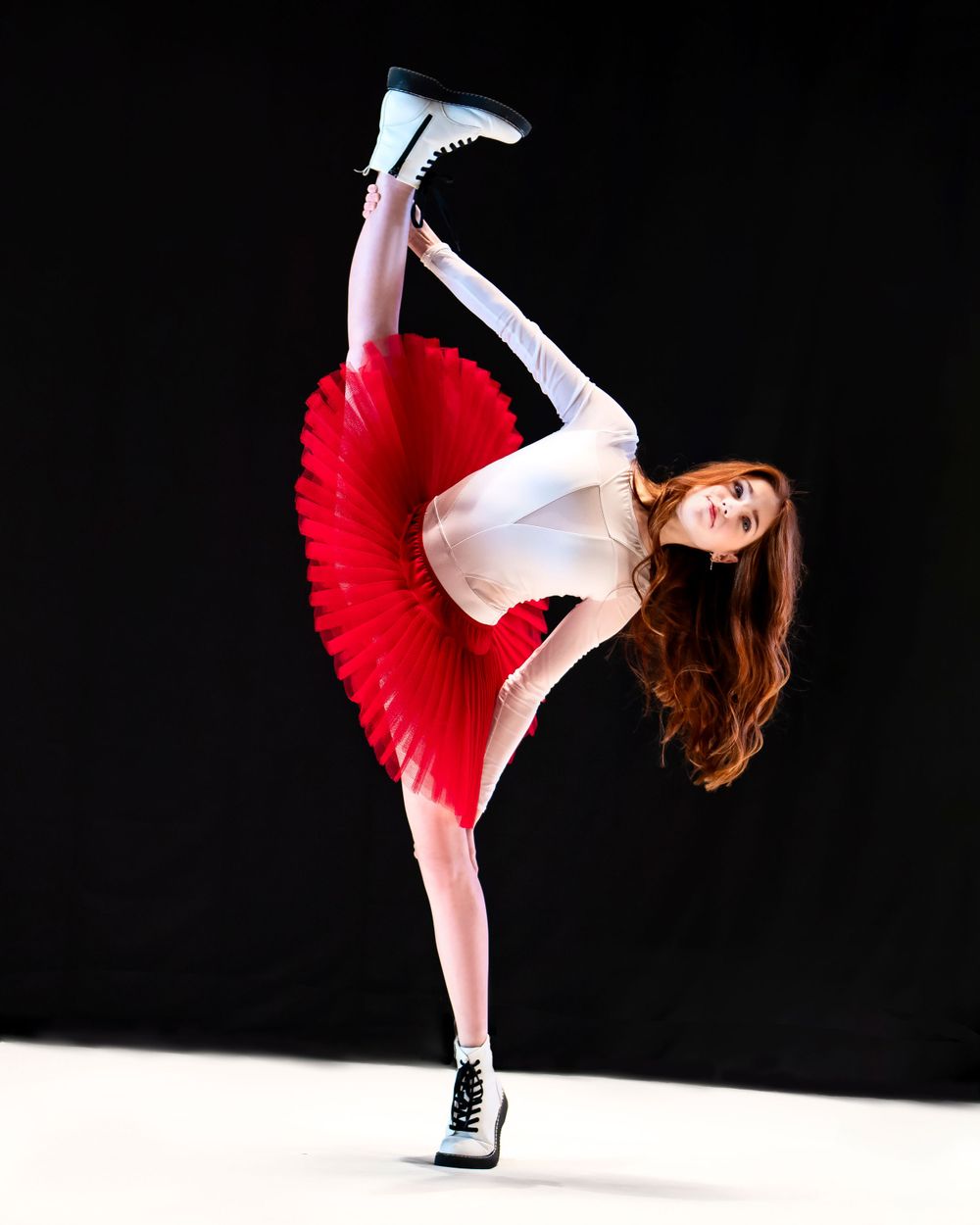 Dancer in a red tutu and white top performs a dramatic high kick pose against a black background.
