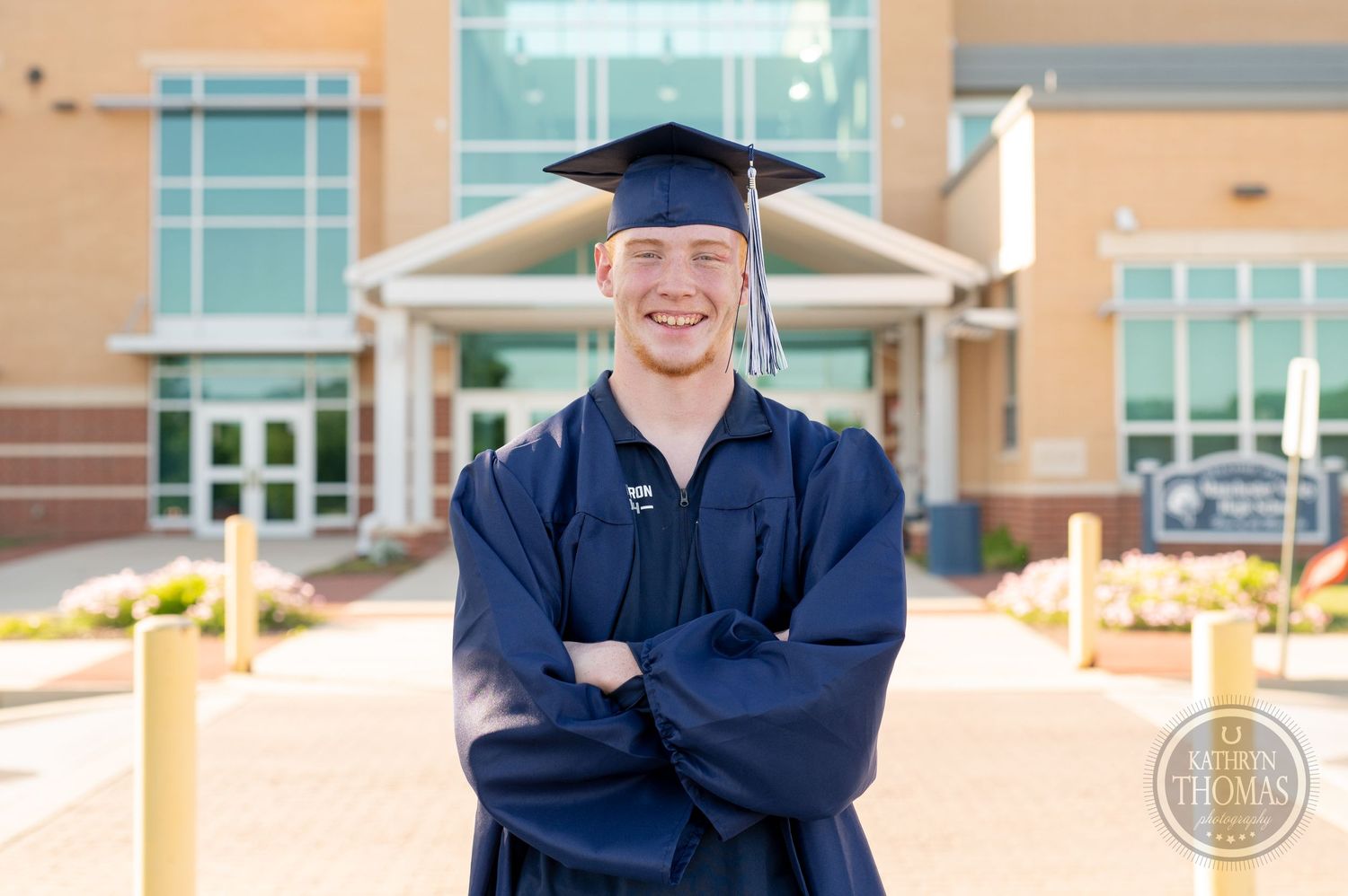 Graduate in traditional commencement regalia poses with crossed arms outside campus building.