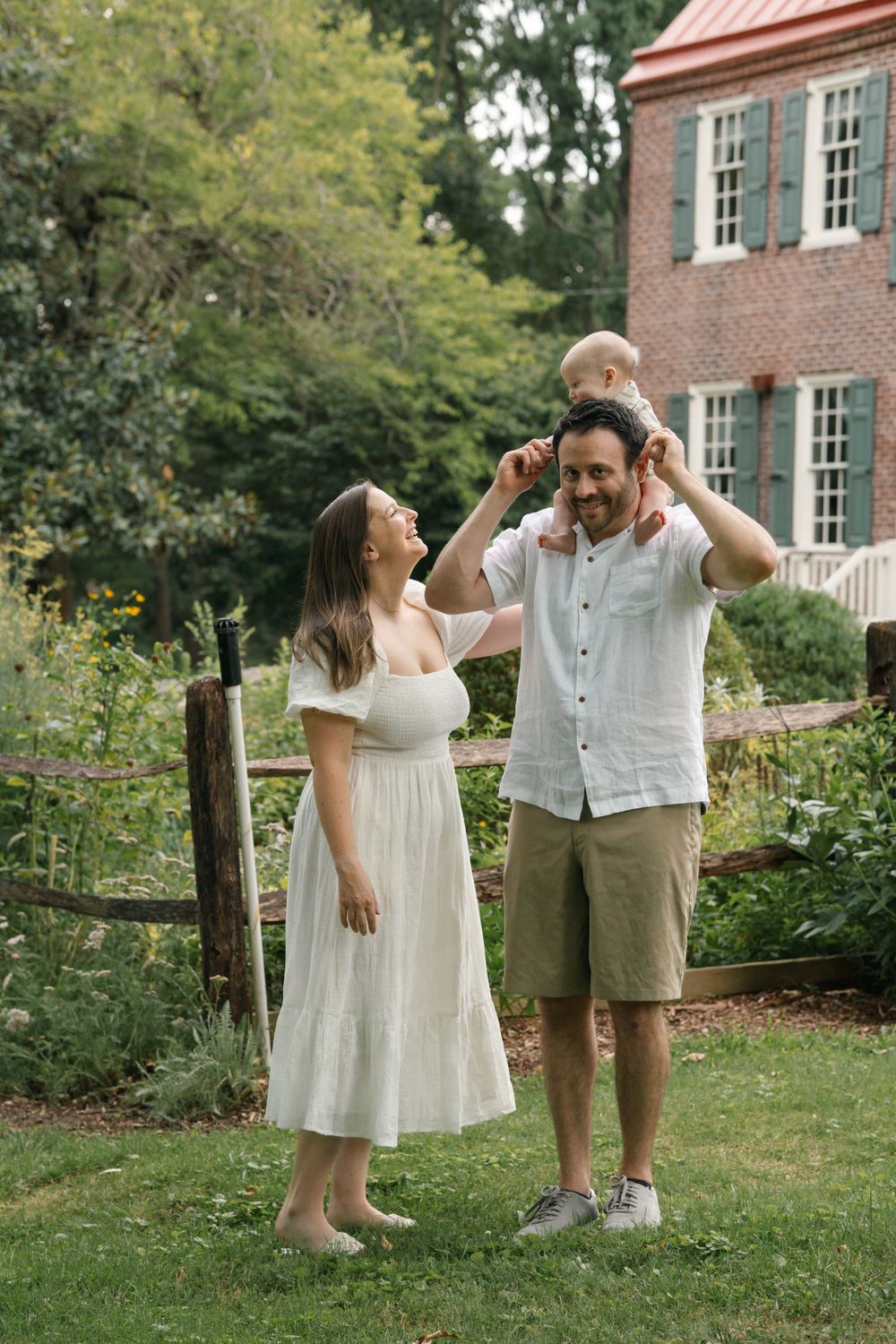 A couple shares a joyful moment with their toddler under overcast skies, with Barclay Farmstead's herb garden in the background.