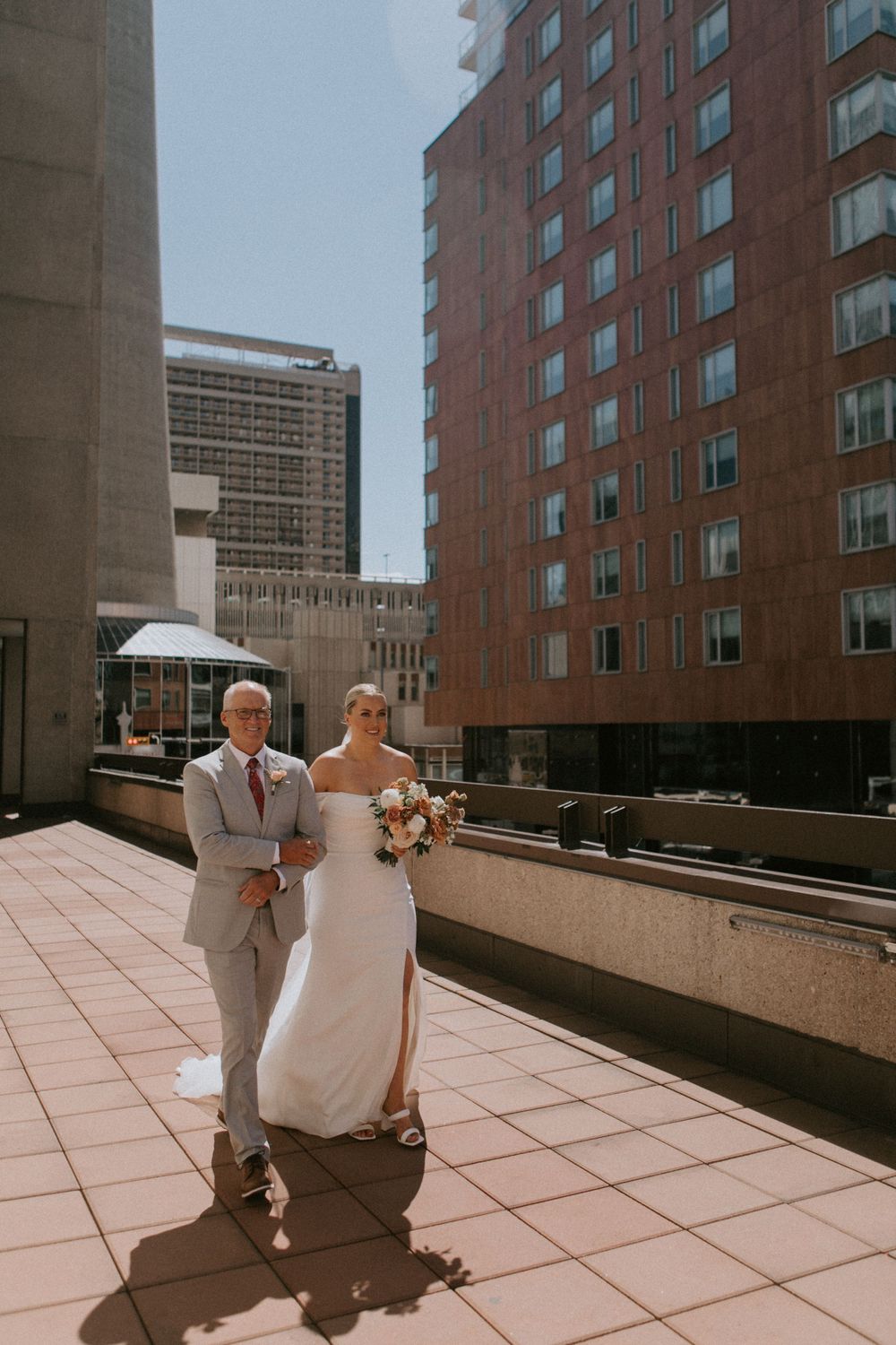 A wedding couple stands on a sunny terrace surrounded by modern city buildings in an urban setting.