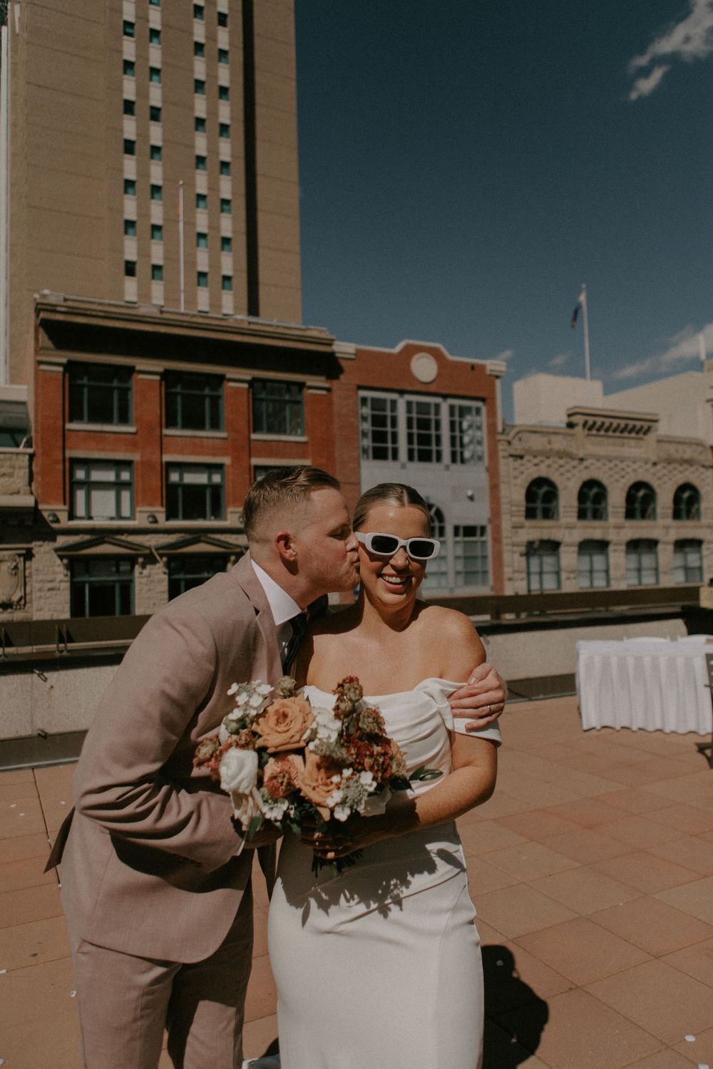 A newlywed couple shares a tender moment on an outdoor terrace with brick buildings and blue sky in the background.