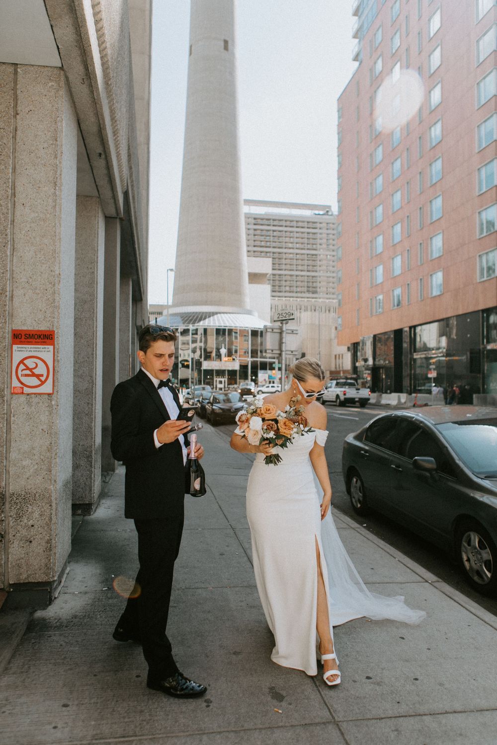A couple in formal wedding attire walks down a city sidewalk between modern buildings holding bouquets.