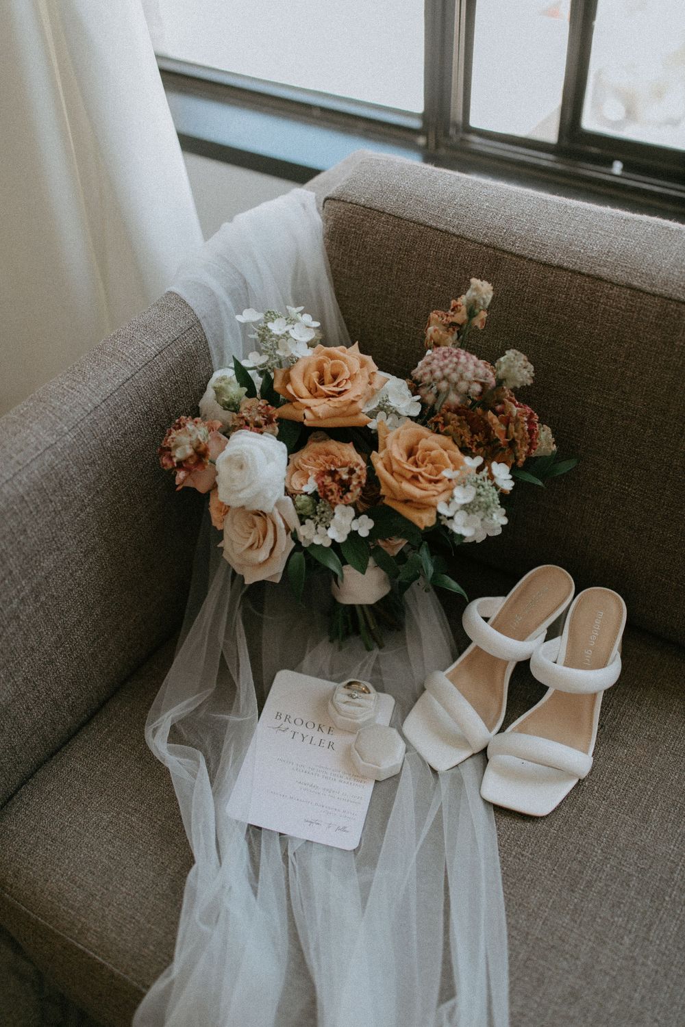 Wedding details with peach and white rose bouquet, white veil, and bridal sandals on a grey couch.