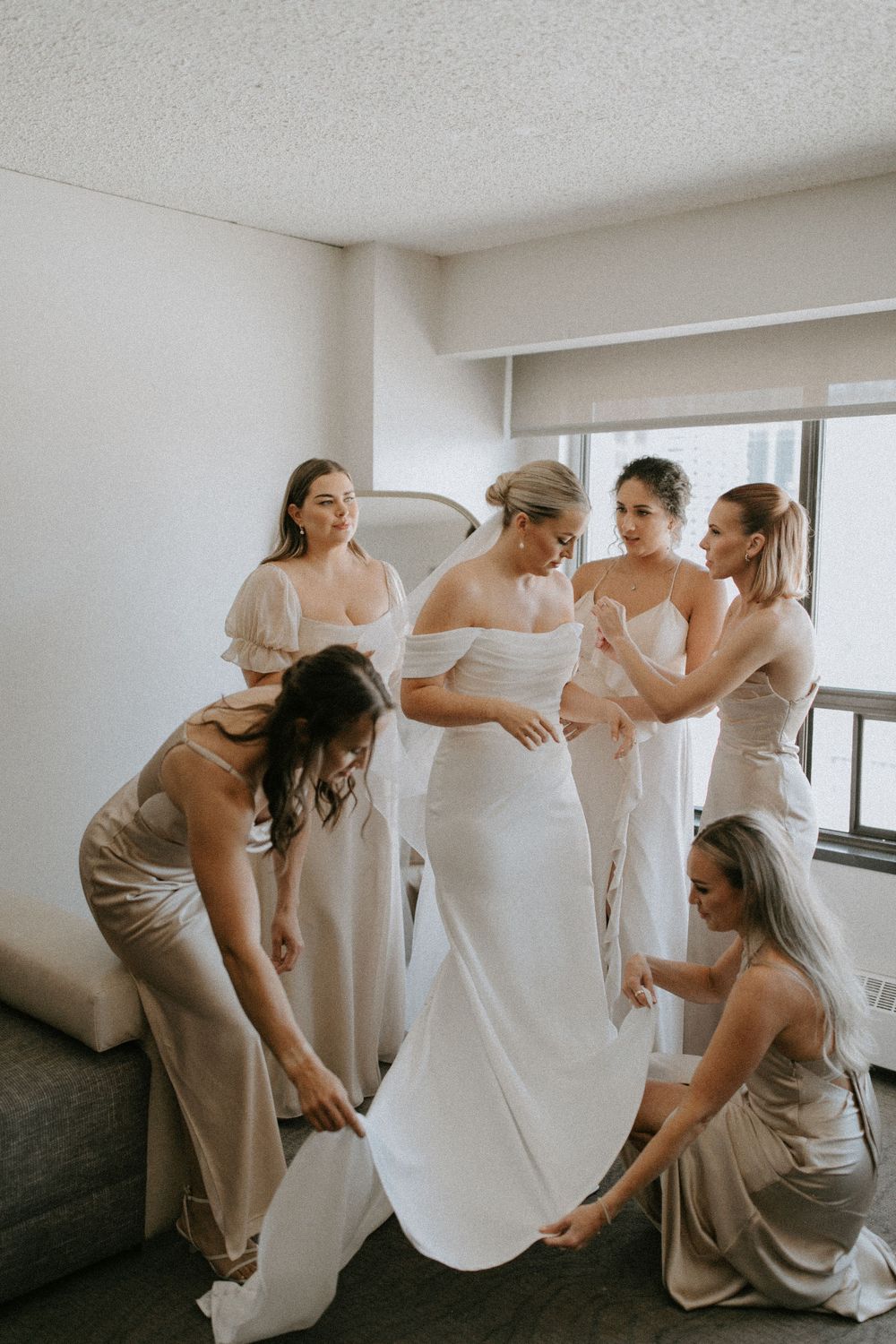 Bridesmaids helping adjust bride's white wedding gown during preparation.