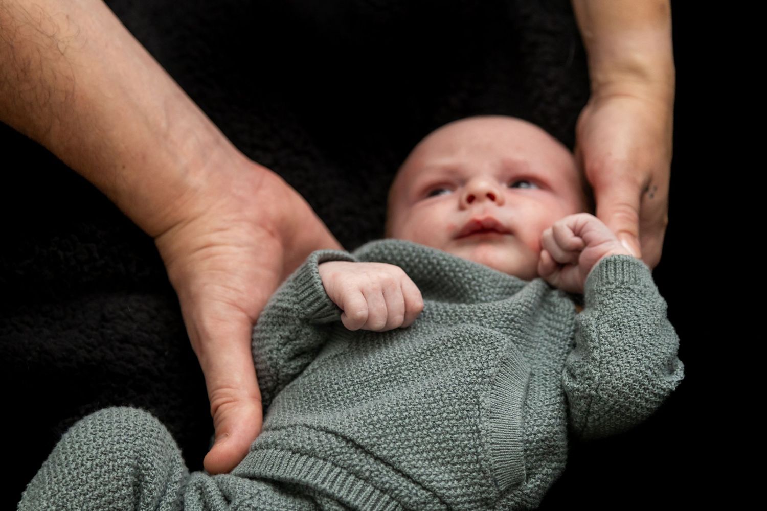 Newborn baby in green knit outfit cradled in adult hands on dark background.