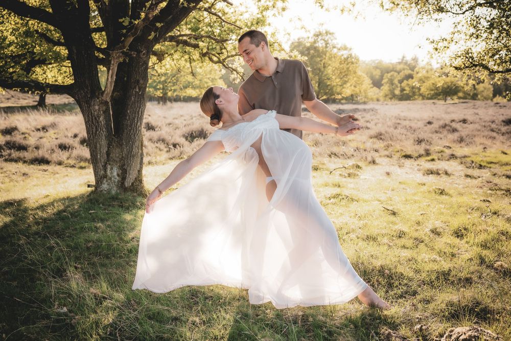 Couple dancing in a golden meadow at sunset with a flowing white wedding dress under a tree.