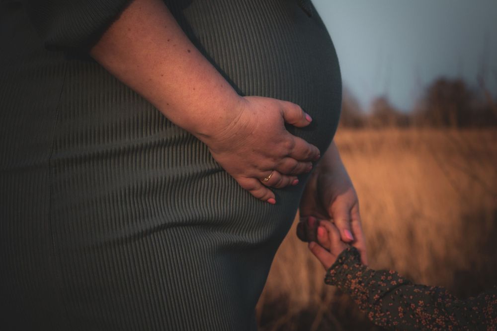 A pregnant woman in a gray dress stands in a golden field at sunset, holding her baby bump tenderly.