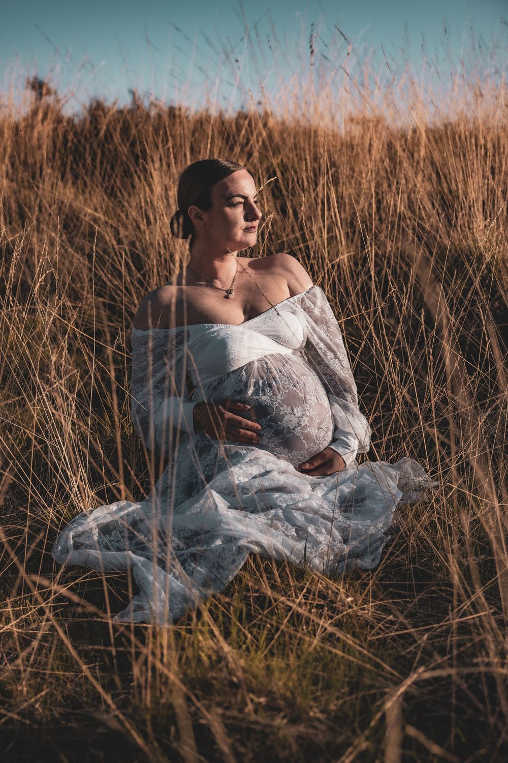 Dreamy maternity photoshoot in a field of tall dried grass at sunset with white flowy dress and natural lighting.
