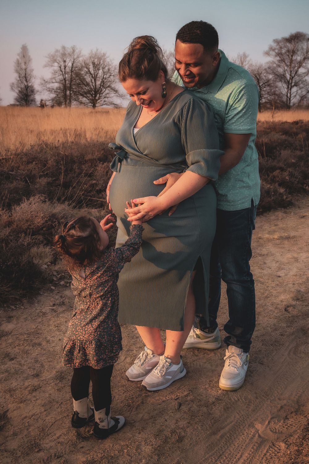A couple and their small child share a tender moment outdoors in a field during golden hour.