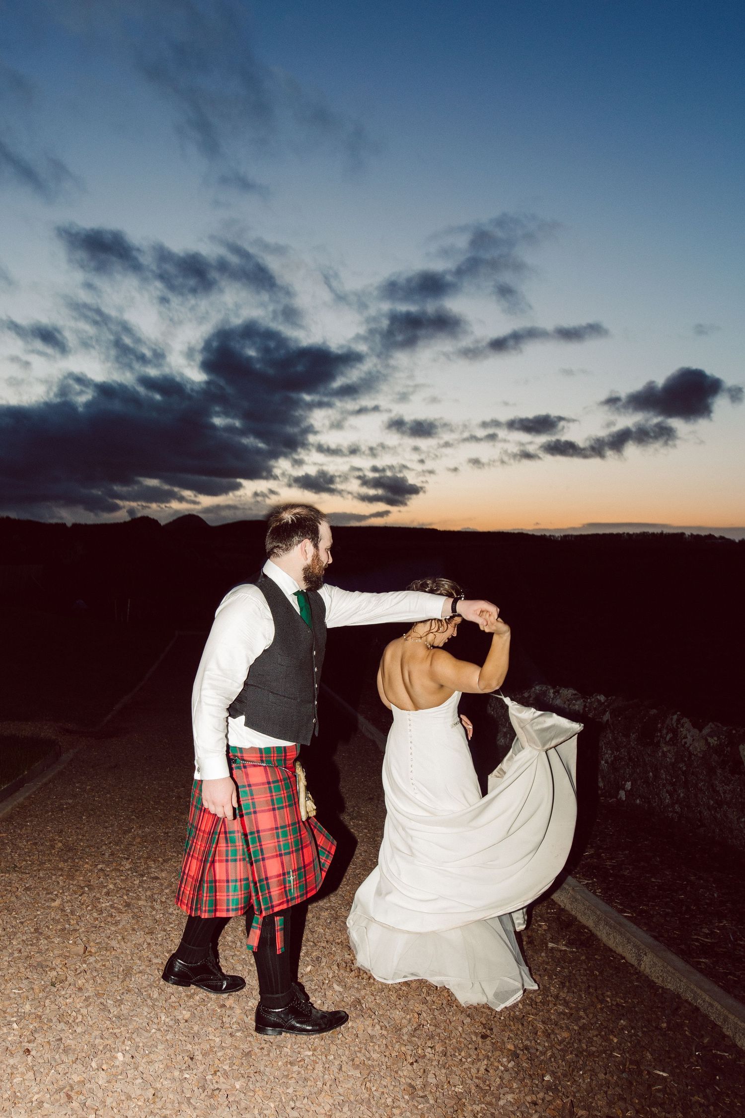 Couple walking at dusk with dramatic sky, one wearing traditional Scottish kilt.